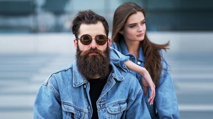 Close Up Portrait von einem Hipster-Paar eines brutalen bärtigen Mannes in Sonnenbrille und seine Freundin in Jeans-Jacken gegen Wolkenkratzer gekleidet.
